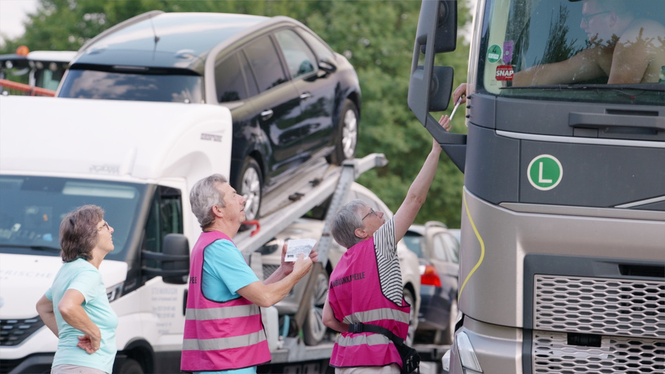 Drei Mitarbeitende der LKW-Seelsorge stehen an einem Lkw. Zwei tragen pinkfarbene Warnwesten, eine weitere Frau ein gleichfarbiges T-Shirt. Eine Mitarbeiterin spricht mit dem Fahrer im Führerhaus. Im Hintergrund stehen ein Autotransporter und weitere Fahr
