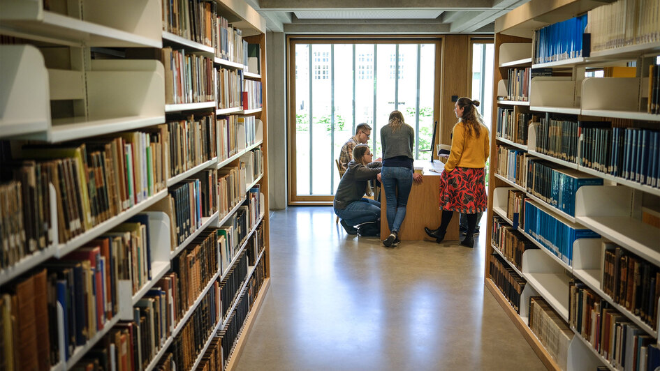 Blick durch einen hellen Gang einer Bibliothek mit hohen, dicht gefüllten Bücherregalen auf beiden Seiten. Am Ende des Gangs stehen und sitzen vier Personen um einen kleinen Tisch vor einer großen Fensterfront.