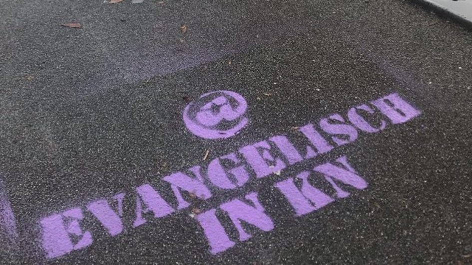 Die Worte @ Evangelisch in KN befinden sich in violetter Schrift auf Asphalt. Die Worte @ Evangelisch in KN befinden sich in violetter Schrift auf Asphalt.