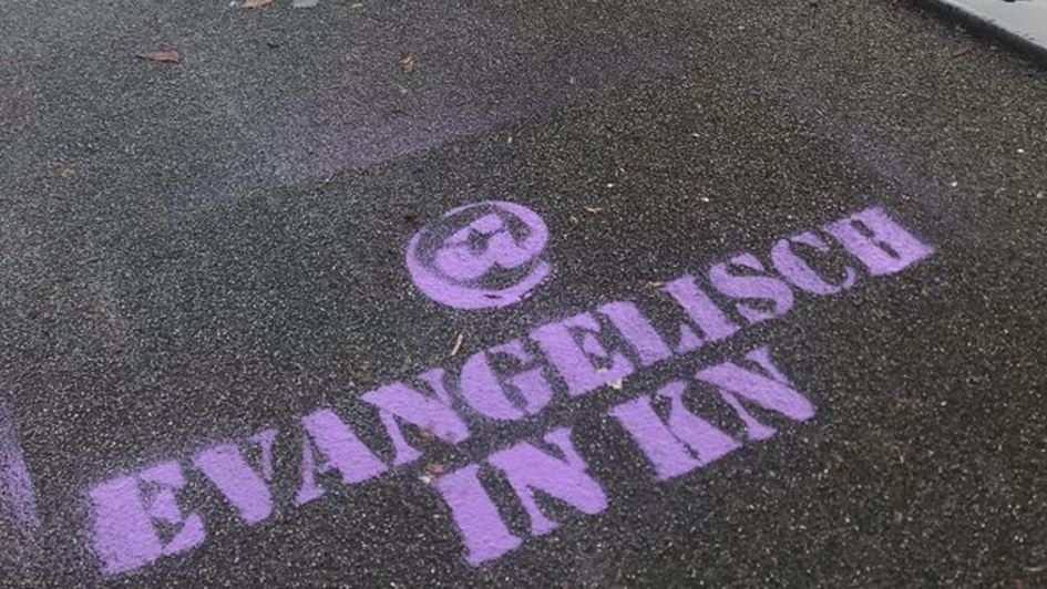 Die Worte @ Evangelisch in KN befinden sich in violetter Schrift auf Asphalt.