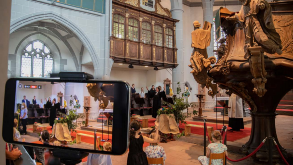 Eine Kirche, in der gerade eine Andacht stattfindet. Im Vordergrund ein Mobilgerät, das den gleichen Raum aufzeichnet. Eine Kirche, in der gerade eine Andacht stattfindet. Im Vordergrund ein Mobilgerät, das den gleichen Raum aufzeichnet.