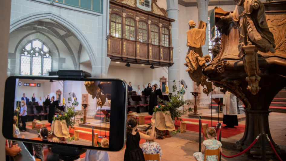 Eine Kirche, in der gerade eine Andacht stattfindet. Im Vordergrund ein Mobilgerät, das den gleichen Raum aufzeichnet.