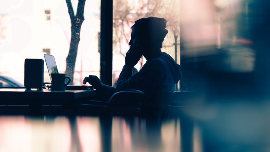 Siluette von einem Menschen an einem Tisch mit Laptop Kaffebecher vor einem großen Fenster.