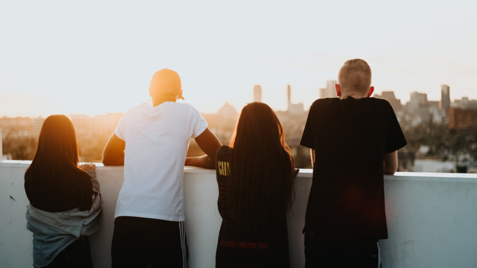 Jugendliche stehen auf dem Dach eines hohen Hauses und blicken auf die Stadt im Sonnenuntergang.