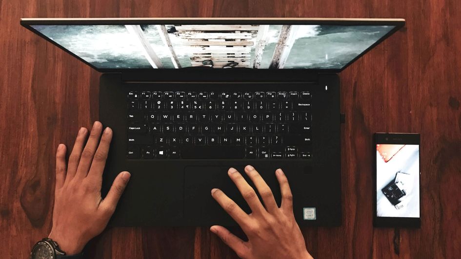 Ein schwarzer geöffneter Laptop von oben, zwei Hände liegen auf der Tastatur. Rechts neben dem Laptop liegt ein Smartphone.