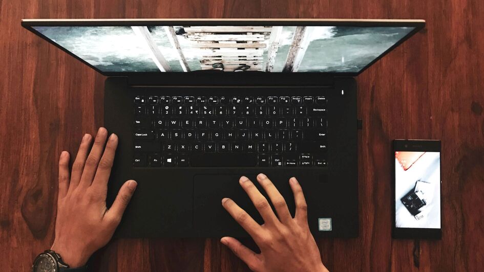 Ein schwarzer geöffneter Laptop von oben, zwei Hände liegen auf der Tastatur. Rechts neben dem Laptop liegt ein Smartphone. Ein schwarzer geöffneter Laptop von oben, zwei Hände liegen auf der Tastatur. Rechts neben dem Laptop liegt ein Smartphone.
