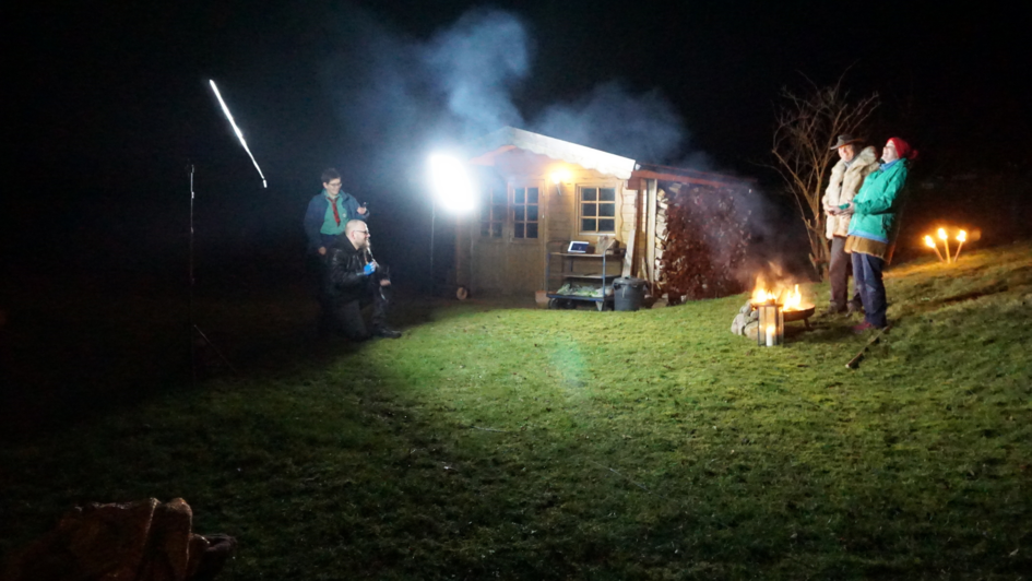 Szene eines Krippenspiels. Vier Menschen stehen und hocken in einem Garten. Es ist dunkel. Licht entsteht durch Scheinwerfer und eine Feuerschale. Im Hintergrund ist einm Gartenhaus zu sehen. Szene eines Krippenspiels. Vier Menschen stehen und hocken in einem Garten. Es ist dunkel. Licht entsteht durch Scheinwerfer und eine Feuerschale. Im Hintergrund ist einm Gartenhaus zu sehen.