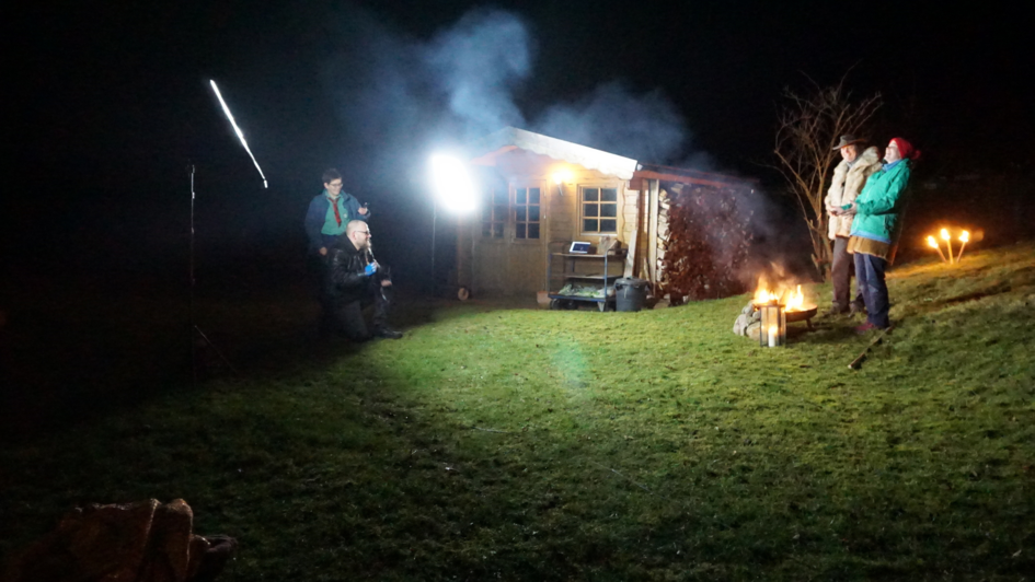 Szene eines Krippenspiels. Vier Menschen stehen und hocken in einem Garten. Es ist dunkel. Licht entsteht durch Scheinwerfer und eine Feuerschale. Im Hintergrund ist einm Gartenhaus zu sehen.