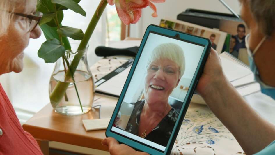 Gesprächssituation mit zwei älteren Menschen über ein Tablet. Unterstützende Person mit OP-Maske hält das Tablet.