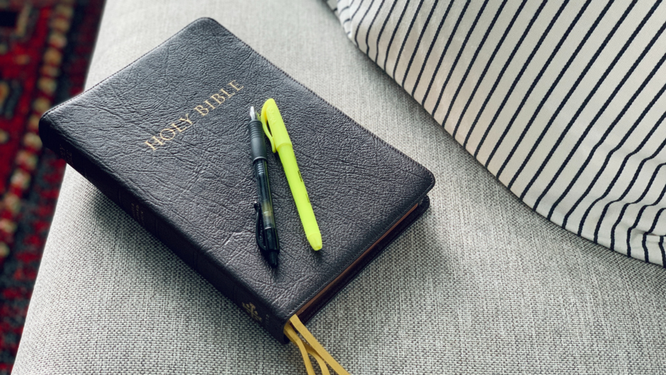 Eine in schwarzem Leder eingebundene Bibel liegt auf einem grauen Sofa. Auf der Bibel liegen zwei Kugelschreiber.  Neben der Bibel liegt ein Kissen mit Streifenmuster.