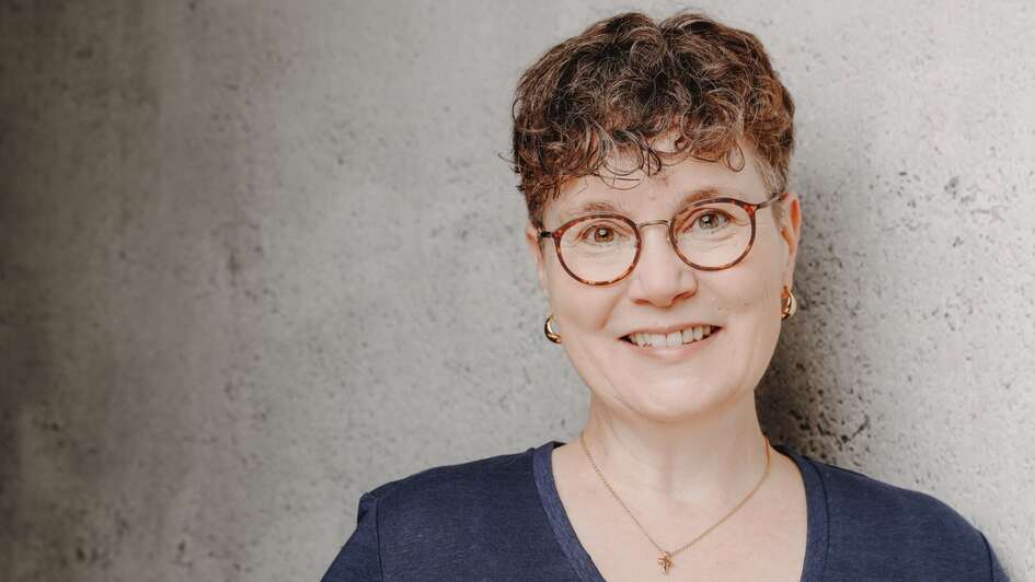 Zu sehen ist die Pfarrerin Svenja Neumann. Sie blickt vor einem grau-steinerener Wand direkt in die Kamera. Svenja Neumann trägt kurze braune Locken und eine Blicke. Sie hat eine goldene Kette an und ein dunkelblaues Oberteil.