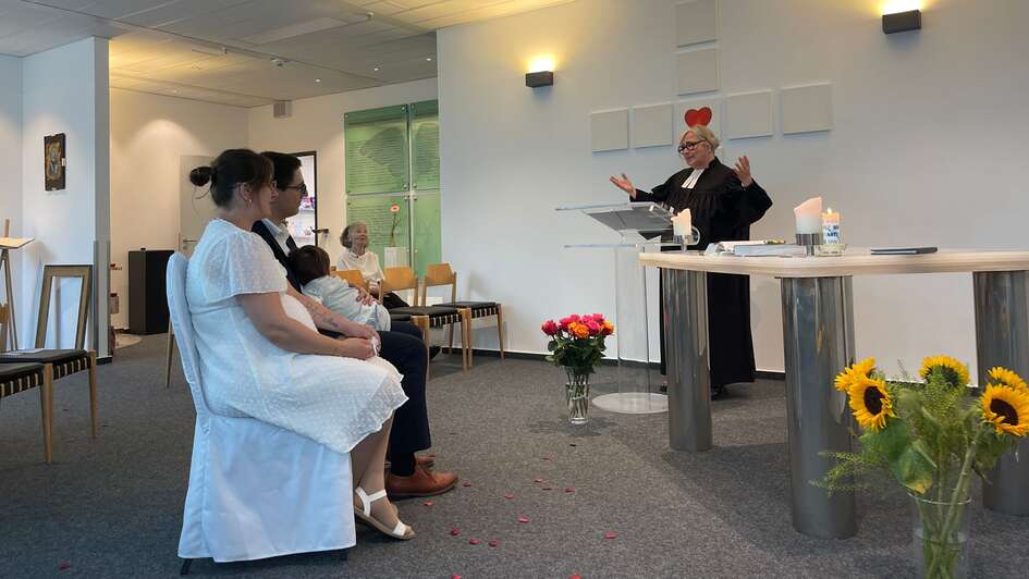 Das Brautpaar Tiffany und Beny sitzt auf den Stühlen, welche mit weißen Hussen überzogen wurden. Auf dem Schoß ihr kleiner Sohn. Pfarrerin Christiane Eßer-Kapp erhebt die Hände zum Segen.
