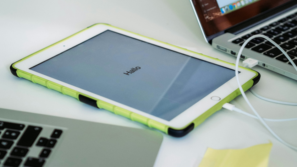 Tablet mit grünem Schutzrahmen liegt auf einem Tisch zwischen zwei Laptops. Auf dem Bildschirm steht das Wort ‚Hallo‘. Ein Ladekabel ist angeschlossen.