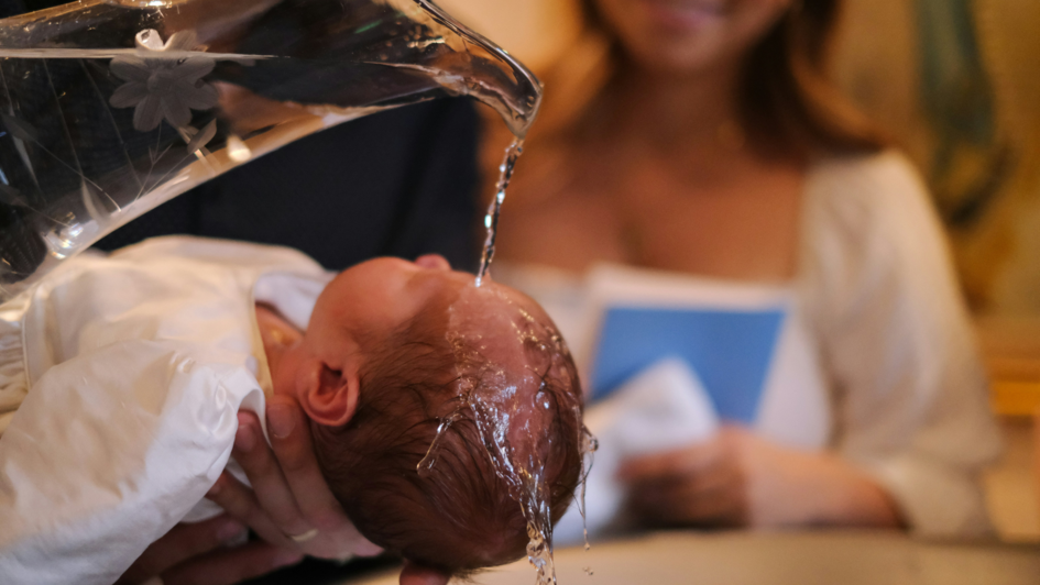 Ein Baby wird bei einer Taufe über einem Taufbecken gehalten, während Wasser aus einer Glaskanne über seinen Kopf gegossen wird. Im Hintergrund hält eine Frau ein blaues Heft und lächelt.