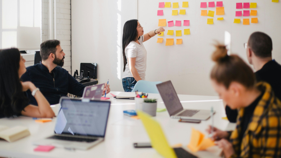 Eine Frau steht an einem Whiteboard mit bunten Haftnotizen und präsentiert einem kleinen Team, das an einem Tisch mit Laptops und Notizmaterialien sitzt. Die Szene zeigt eine kreative Arbeits- oder Diskussionssituation in einem hellen, modernen Büro.