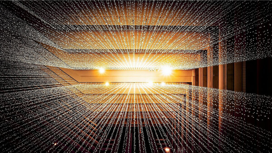 Installation aus Tausenden kleiner Lichtpunkte, die in geraden Linien von der Decke hängen und in der Perspektive wie ein strahlenförmiger Lichttunnel wirken; warme, goldene Lichtstimmung in einem mehrstöckigen Innenraum.
