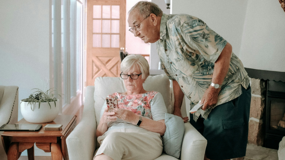 Älteres Paar in einem Wohnzimmer: Eine Frau sitzt auf einem Sessel und blickt konzentriert auf ihr Smartphone, während ein Mann sich über sie beugt und mit Interesse auf den Bildschirm schaut. Neben ihr liegt ein Tablet auf einem Beistelltisch. Älteres Paar in einem Wohnzimmer: Eine Frau sitzt auf einem Sessel und blickt konzentriert auf ihr Smartphone, während ein Mann sich über sie beugt und mit Interesse auf den Bildschirm schaut. Neben ihr liegt ein Tablet auf einem Beistelltisch.