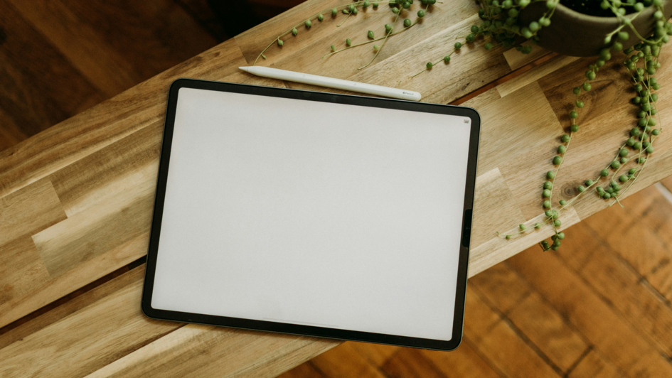 Tablet mit weißem Bildschirm und Stylus auf einem Holztisch neben einer hängenden Grünpflanze – aus der Vogelperspektive fotografiert. Tablet mit weißem Bildschirm und Stylus auf einem Holztisch neben einer hängenden Grünpflanze – aus der Vogelperspektive fotografiert.