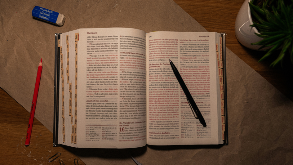 Aufgeschlagene Bibel auf einem Schreibtisch mit Stift, Radiergummi, Bleistift und Büroklammern – Symbol für theologisches Lernen und Studium.