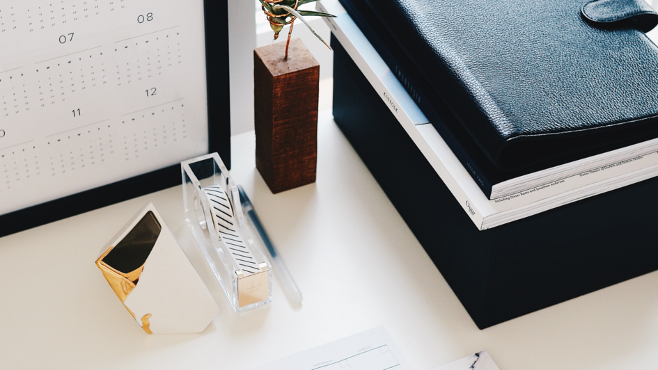 Auf einem Schreibtisch liegen mehrere, teilweise in Leder eingeschlagene Notizbücher und Hefte. An der Wand ist ein Kalender mit einem schwarzen Rahmen angelehnt. Davor befinden sich ein Klebebandspender und ein Briefbeschwerer. Neben dem Kalender befin Auf einem Schreibtisch liegen mehrere, teilweise in Leder eingeschlagene Notizbücher und Hefte. An der Wand ist ein Kalender mit einem schwarzen Rahmen angelehnt. Davor befinden sich ein Klebebandspender und ein Briefbeschwerer. Neben dem Kalender befin