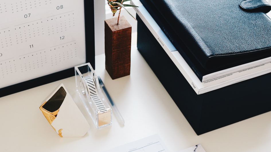 Auf einem Schreibtisch liegen mehrere, teilweise in Leder eingeschlagene Notizbücher und Hefte.  An der Wand ist ein Kalender mit einem schwarzen Rahmen angelehnt. Davor befinden sich ein Klebebandspender und ein Briefbeschwerer.  Neben dem Kalender befin