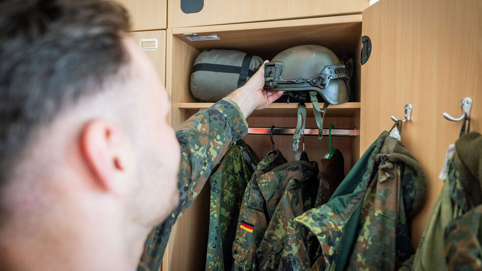 Wehrpflicht: Ein Bundeswehrsoldat in Flecktarnuniform greift in einem geöffneten Holzspind nach einem olivgrünen Schutzhelm. Im Spind hängen weitere Uniformteile und militärische Ausrüstung.