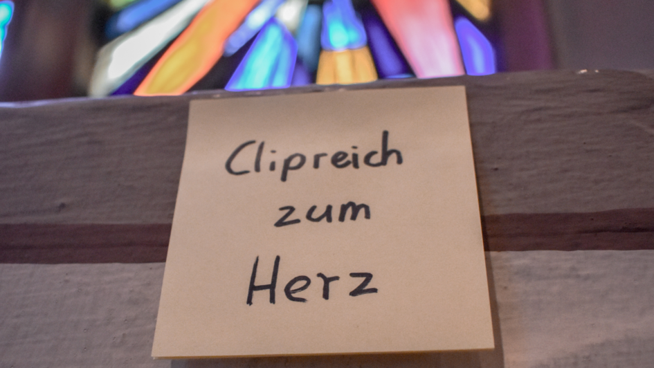 Ein handgeschriebenes Post-it mit der Aufschrift „Clipreich zum Herz“ ist an eine Holzfläche geheftet. Im Hintergrund sind bunte Kirchenfenster zu sehen, die in verschiedenen Farben leuchten.