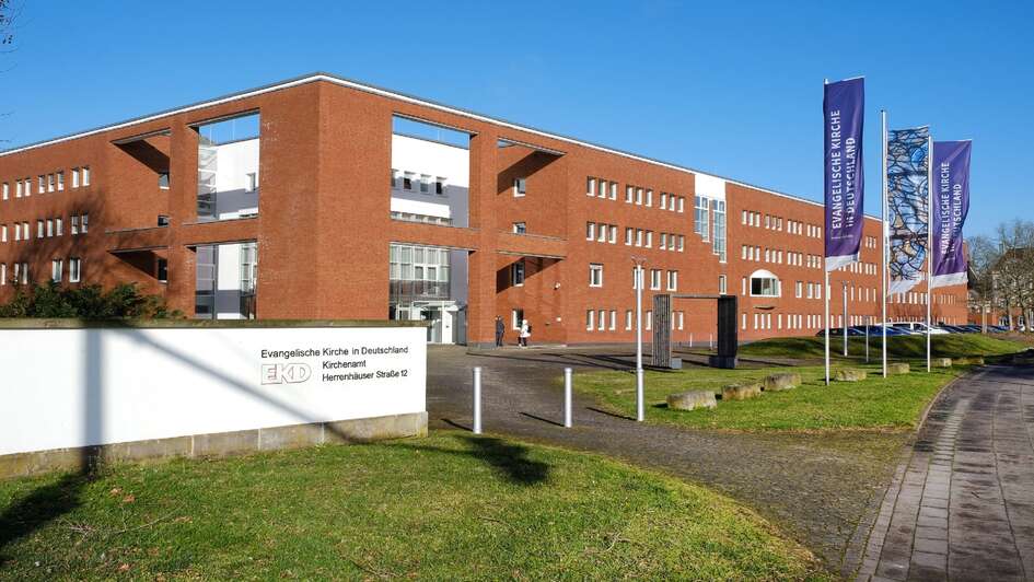 Zu sehen ist das rote Backsteingebauede der EKD in Hannover. Der Himmel ist blau. Vor dem rechteckigen Gebauede wehen lila Fahnen mit dem EKD Logo