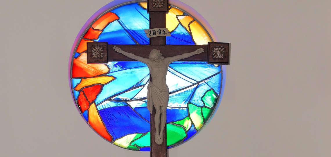 Kreuz vor farbigem, runden Kirchenfenster