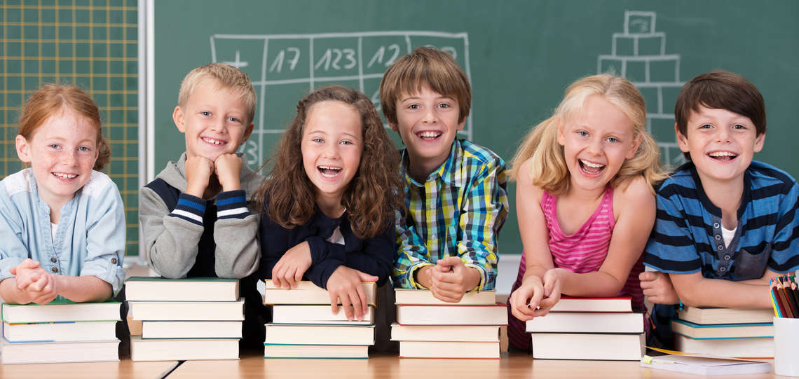 Lachende Kinder mit Stapeln an Büchern vor einer Schultafel