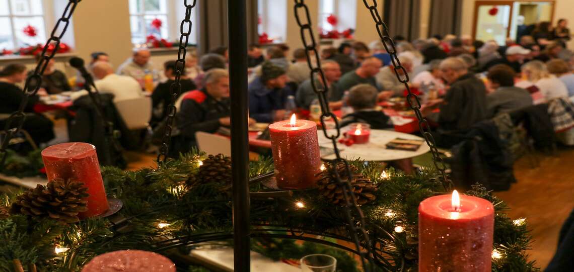Im Vordergrun steht ein Adventskranz. Zwei Kerzen brennen. verschwommen im Hintergrund sind Tischreihen zu sehen. Menschen sitzen daran und essen. Der Saal ist voll