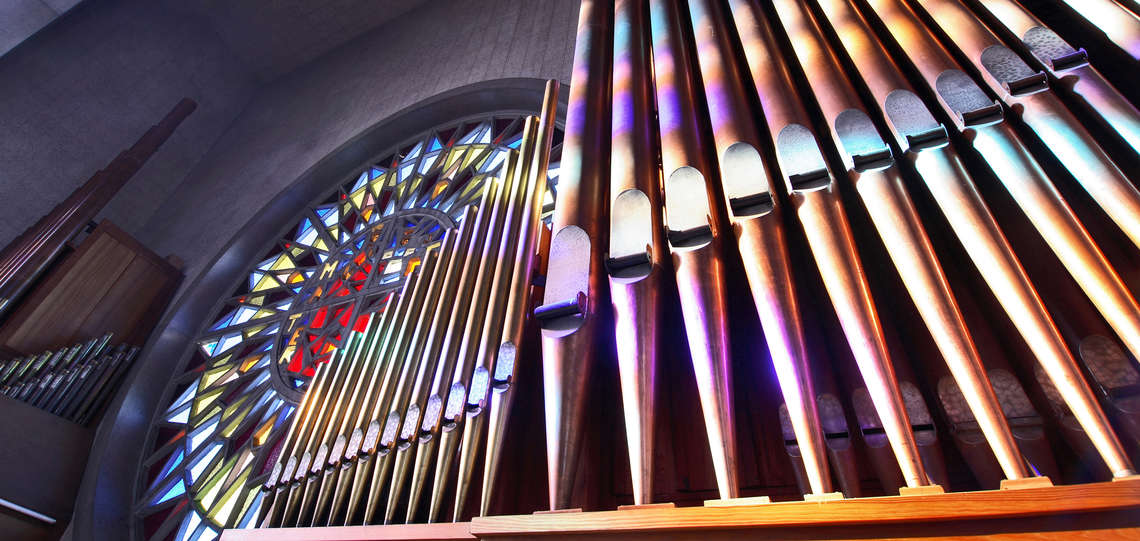 Orgelpfeifen neben einem Buntglasfenster in einer Kirche.