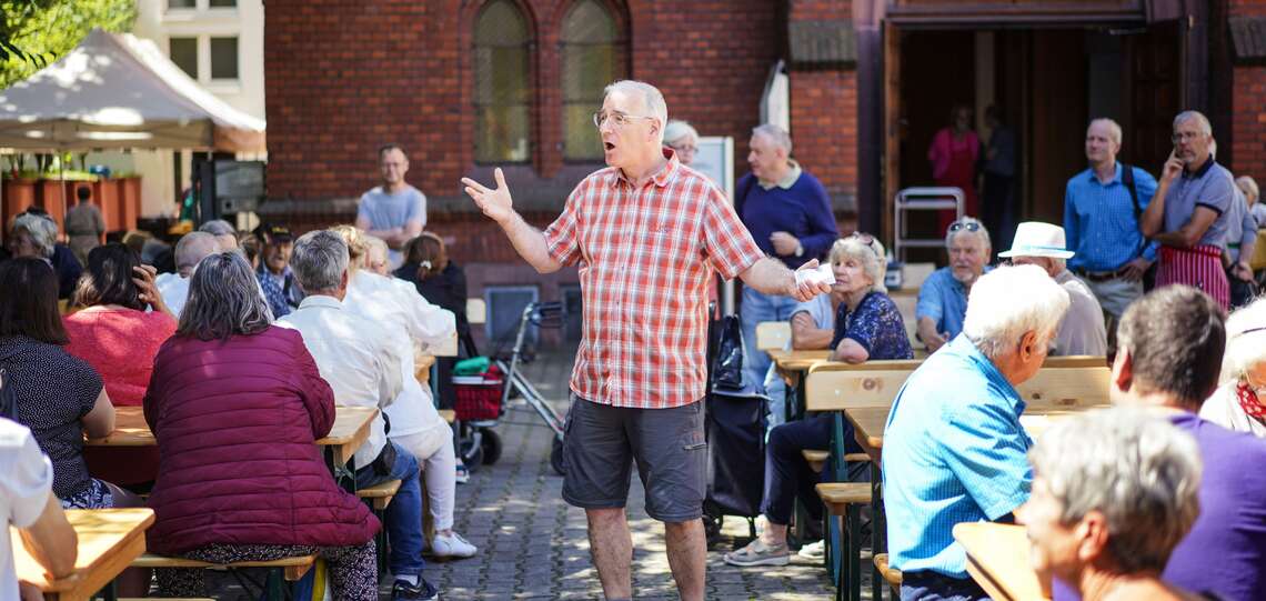 Pastor Uwe Sassnowski (Saßnowski) beim "Mittagstisch fuer alle" an der Evangelisch-methodistischen Christuskirche am Merianplatz in Frankfurt am Main Um ihn herum sitzen Menschen an Bierzeltgarnituren und essen ihr Mittagessen.