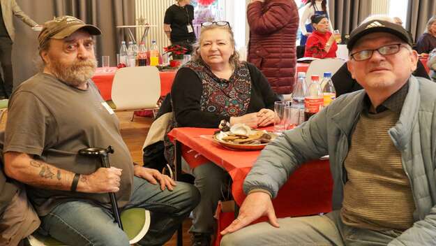 Zu sehen sind links ein Mann über 50 mit Bart und Kappe rechts im Bild sitzend. Neben ihm seine Frau. Auf der rechten Seite sicht ein weiterer Heer, mit Brille und Kappe. Er blickt in die Kamera.