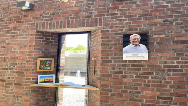 Zu sehen ist ein Fenster in der Autobahnkirche Medenbach. Davor liegt das Andachtsbuch. An der rechten Wand haengt ein Foto des Stifters der Kirche Alfred Weigle