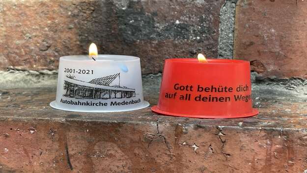 zwei brennende Kerzen, etwas groesser als ein Teelicht. Die links Kerze steht in weißer Plastikhuelle. Darauf die Aufschrift Medenbach 2001-2021. Die rechte Kerze steht in einer roten Plastikhuelle. Darauf die Aufschrift: Gott behuete dich auf all deinen 