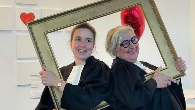 Die beiden Pfarrerinnen Mirjam Biskamp_Krämer und Christiane Eßer-Kapp im Talar in der Flughafenkapelle stehend. Sie blicken durch einen hölzernen großen Bilderrahmen in die Kamera
