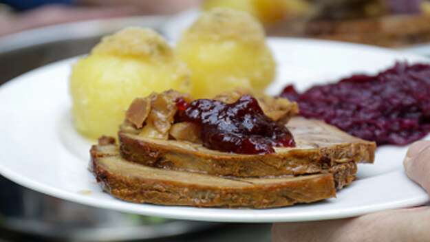 Zu sehen ist ein weißer Teller. Darauf liegen im Hintergrund unscharf Kartoffelklöße. Im Vordergrund ist eine Schreibe Weihnachtsbraten mit Preiselbeeren zu sehen. 