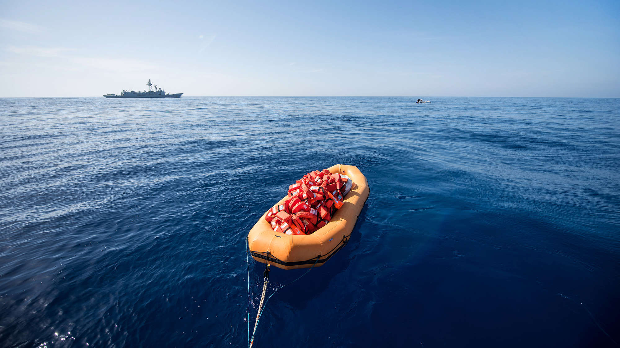 Ein Schlauchboot der Sea Watch-2 auf dem die Rettungswesten zu den Schlauchbooten der Geflüchteten transportiert werden. Im Hintergrund links ein spanisches Kriegsschiff der Frontex-Mission. Es hat die Rettung der Menschen im Schlauchboot hinten rechts be