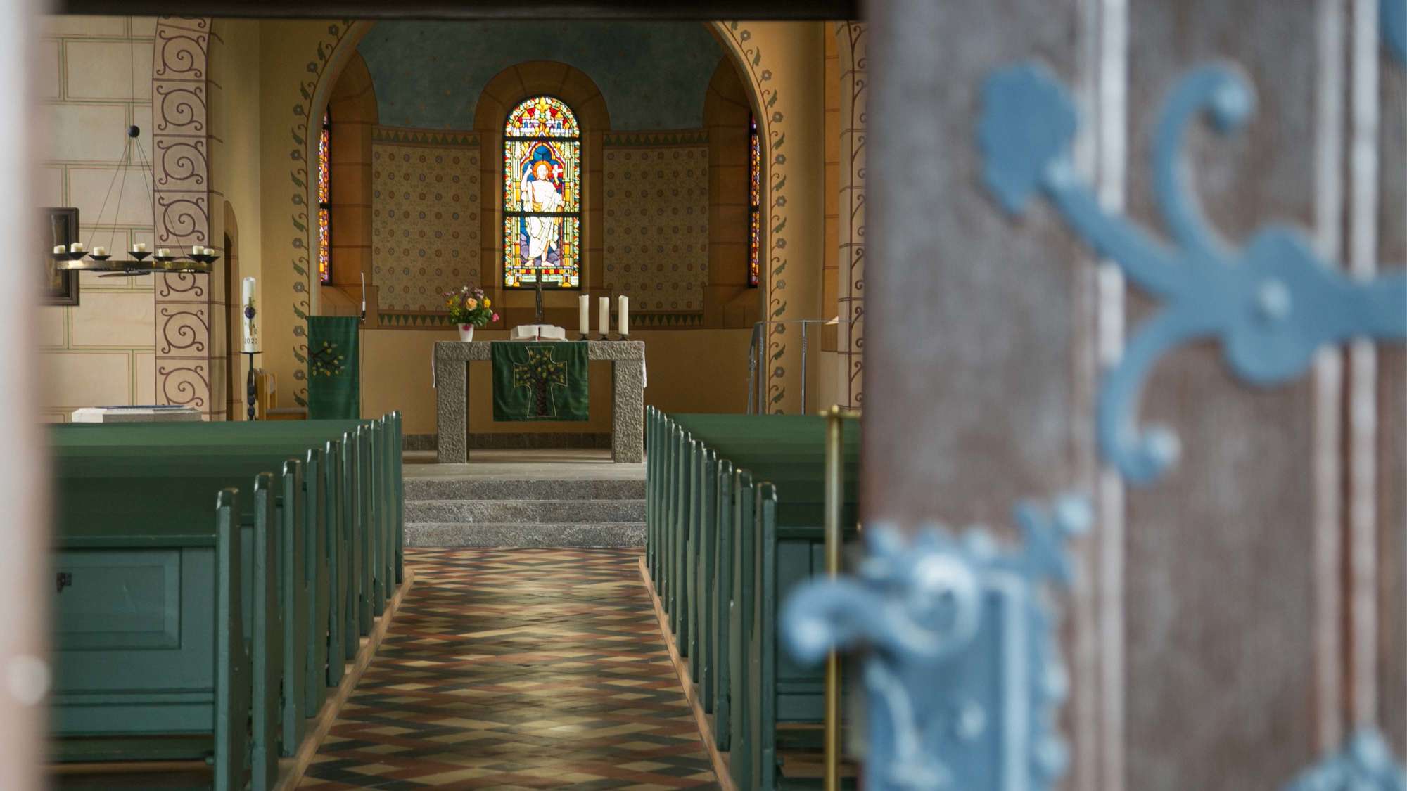 Im Vordergrund rechts die Eingangstür zur Kirche. Bunte Bodenfliesen, Verzierungen am Bogen. Helle Fenster, im Altarraum hellgrauer Steinboden.
